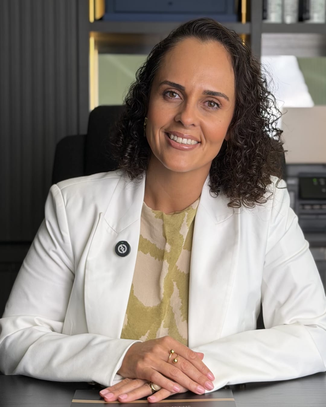 Francine Sinnott sorrindo, de blazer branco, com as mãos sobre a mesa.